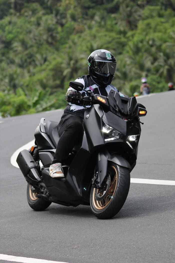 Motorcyclist in full gear rides a sleek black scooter on a winding road through lush greenery.